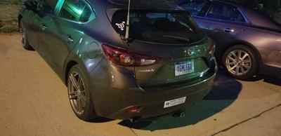 A 2014 Mazda3 hatchback with a large ham radio antenna on the hatch, with a new white transgender logo with the center circle filled with the biohazard symbol
