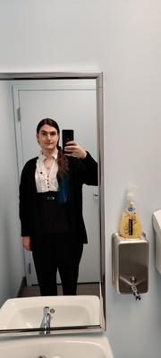 A solemn woman, clearly close to crying, takes a selfie in an office mirror. She is wearing her long dark hair in a ponytail over her right shoulder, showing that the lower third is dyed deep blue. She has a black cardigan on over a dressy white and black blouse, and black dress slacks. A She/Her pronoun pin is on her lapel.